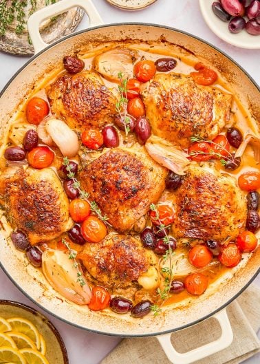 A skillet with roasted chicken thighs, cherry tomatoes, shallots, olives, and herbs in a savory sauce.