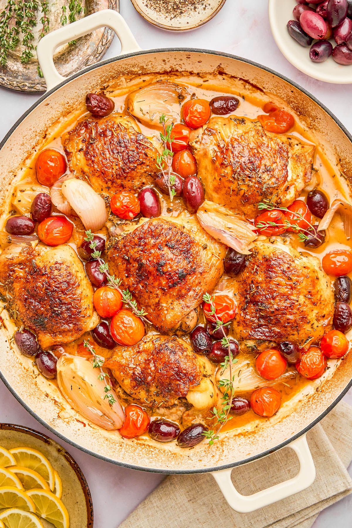 A skillet with roasted chicken thighs, cherry tomatoes, shallots, olives, and herbs in a savory sauce.