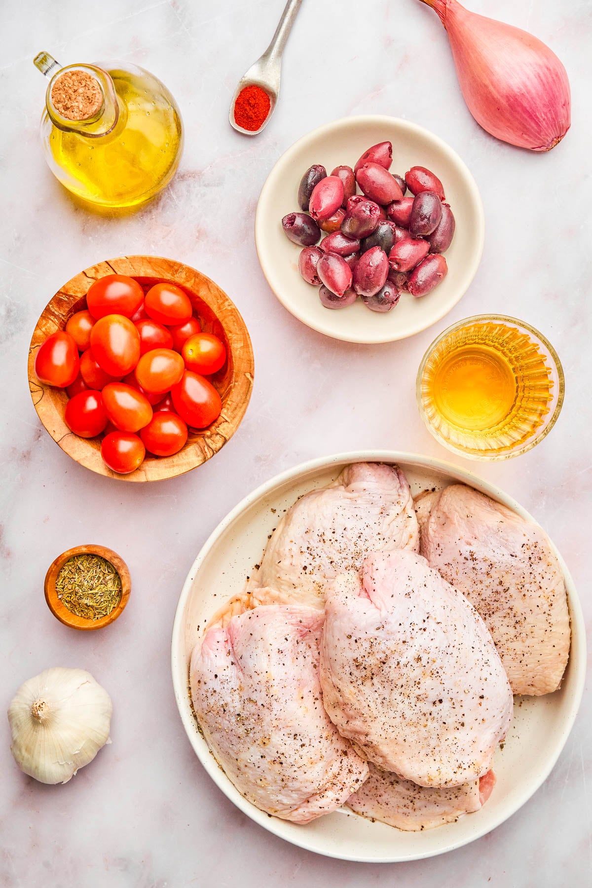 Raw chicken thighs on a plate, surrounded by cherry tomatoes, Kalamata olives, a shallot, garlic, olive oil, white wine, dried herbs, and a spoon of paprika on a white surface.