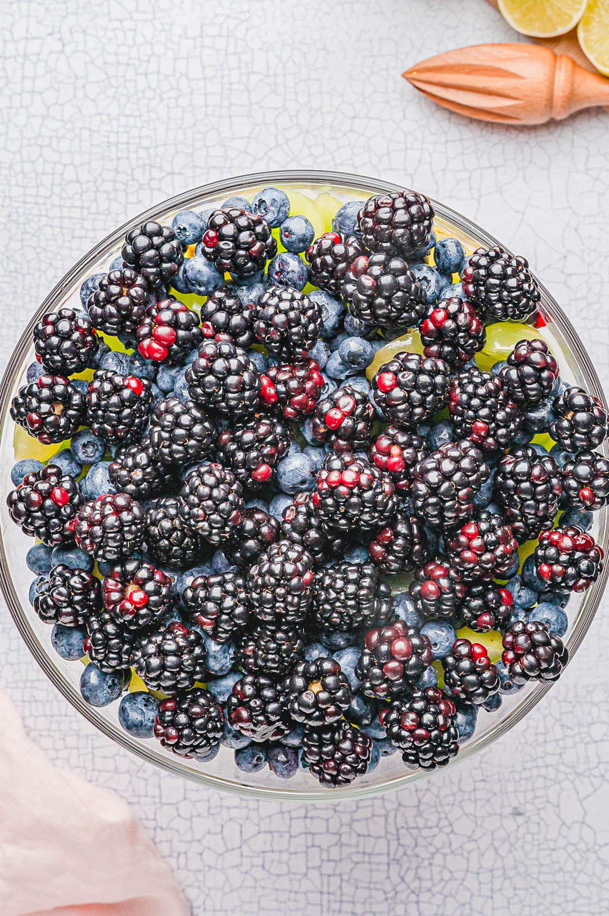 A glass bowl filled with fresh blackberries and blueberries, placed on a light textured surface with a citrus juicer and halved lime in the background.