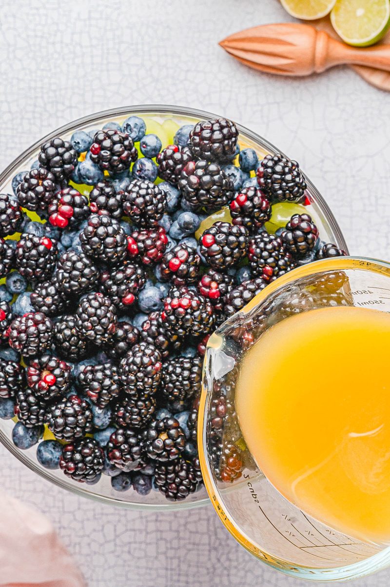 A glass bowl filled with fresh blackberries and blueberries, with a measuring cup of orange juice being poured in.