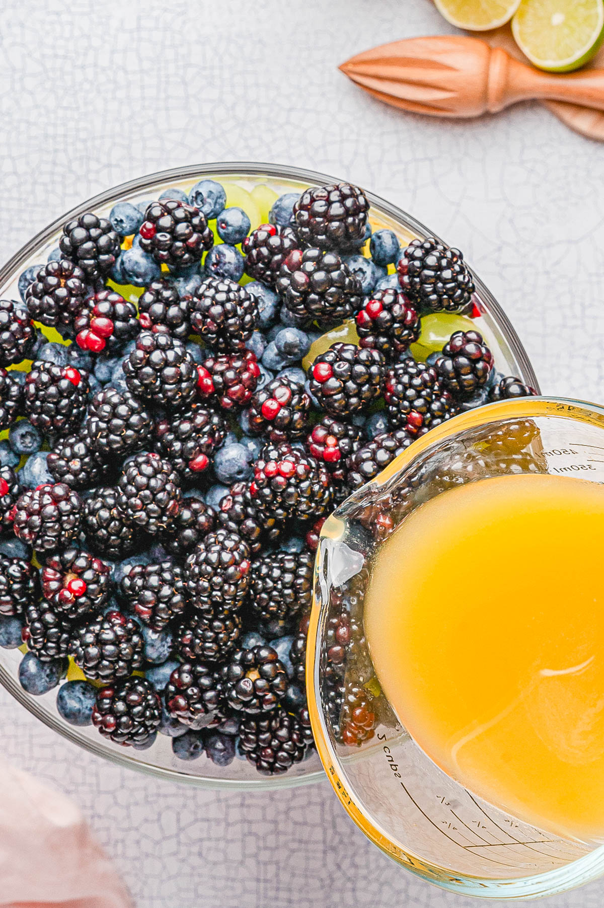 A glass bowl filled with fresh blackberries and blueberries, with a measuring cup of orange juice being poured in.