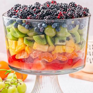 A large glass trifle dish filled with layers of assorted fresh fruits, including berries, kiwi, grapes, pineapple, orange slices, and strawberries.