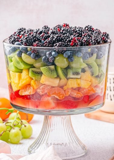 A large glass trifle dish filled with layers of assorted fresh fruits, including berries, kiwi, grapes, pineapple, orange slices, and strawberries.