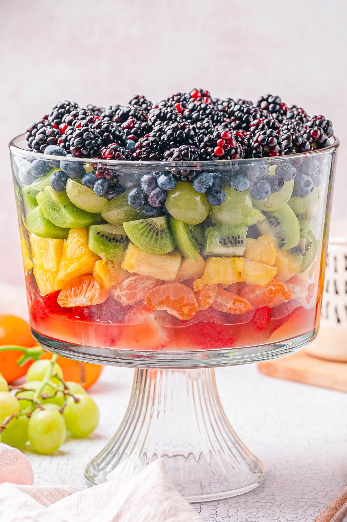 A large glass trifle dish filled with layers of assorted fresh fruits, including berries, kiwi, grapes, pineapple, orange slices, and strawberries.