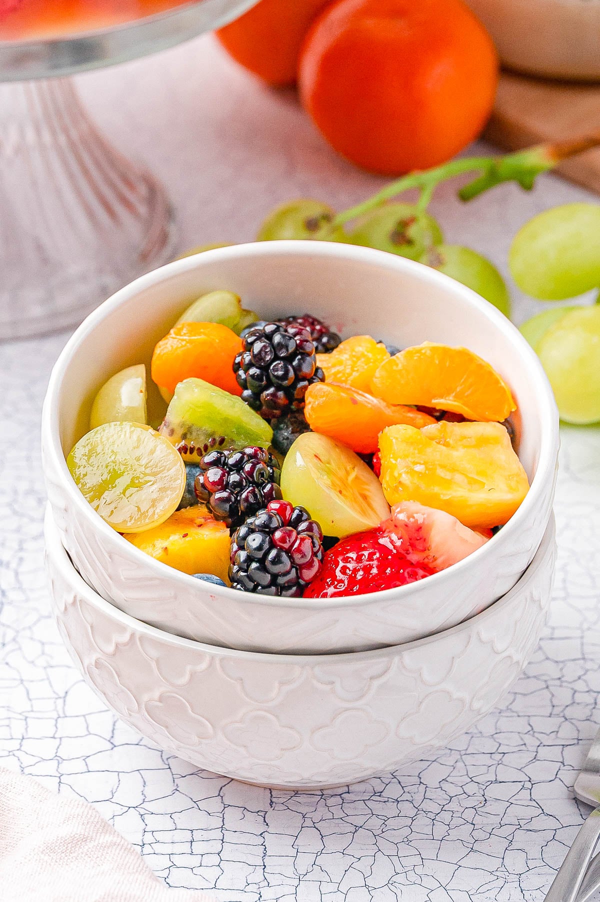 A white bowl filled with a mixed fruit salad, including blackberries, grapes, mandarin oranges, pineapple, and strawberries, sits on a cracked white surface.