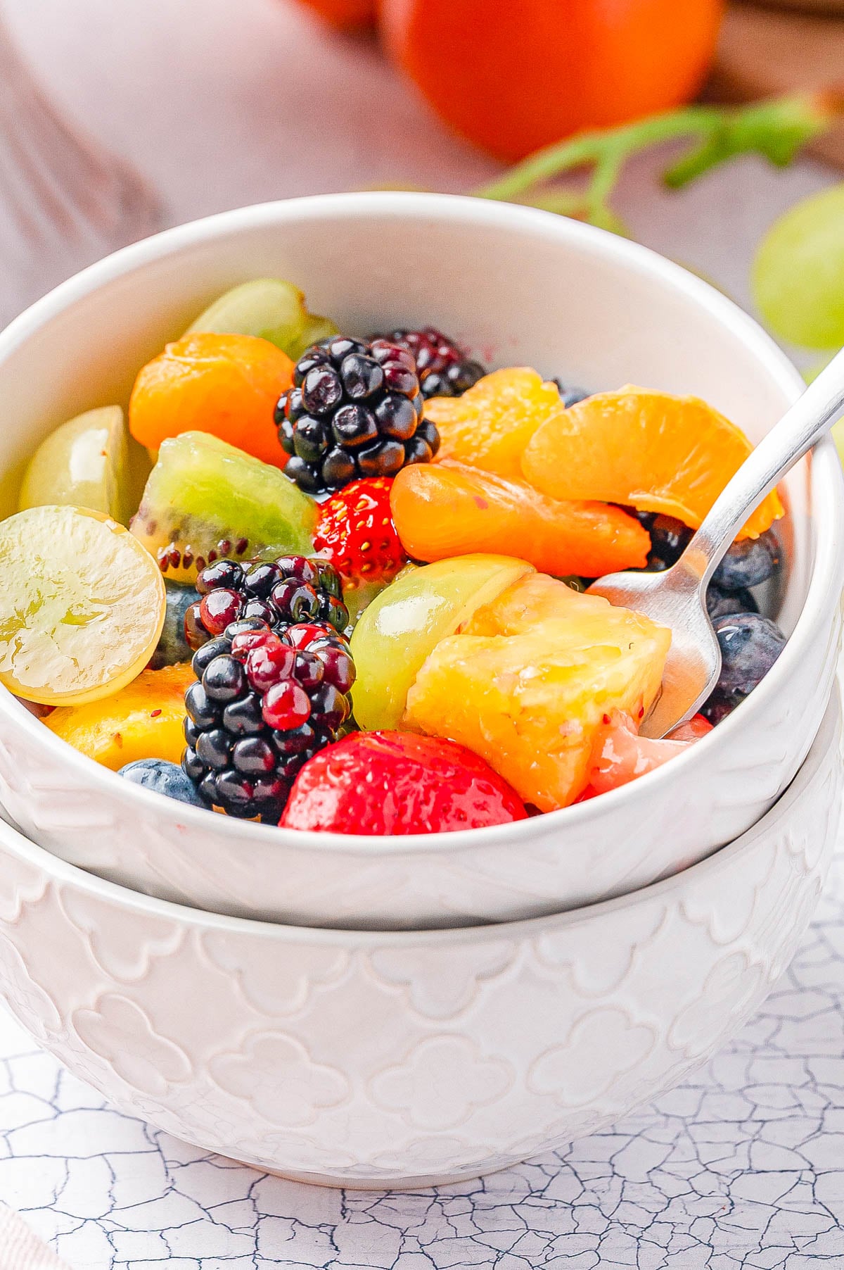 A white bowl filled with assorted fresh fruit, including blackberries, strawberries, grapes, blueberries, mandarin oranges, and pineapple, with a spoon resting inside.