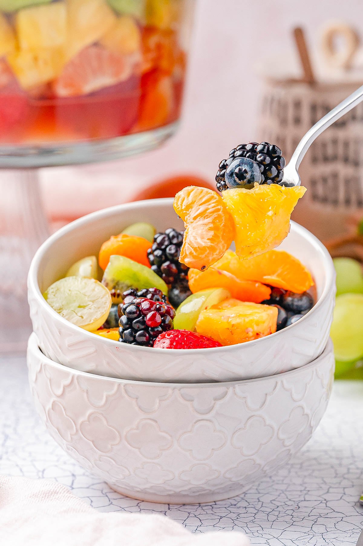 A white bowl filled with fresh fruit salad, including blackberries, mandarin orange segments, grapes, and strawberries, with a spoon lifting a portion.