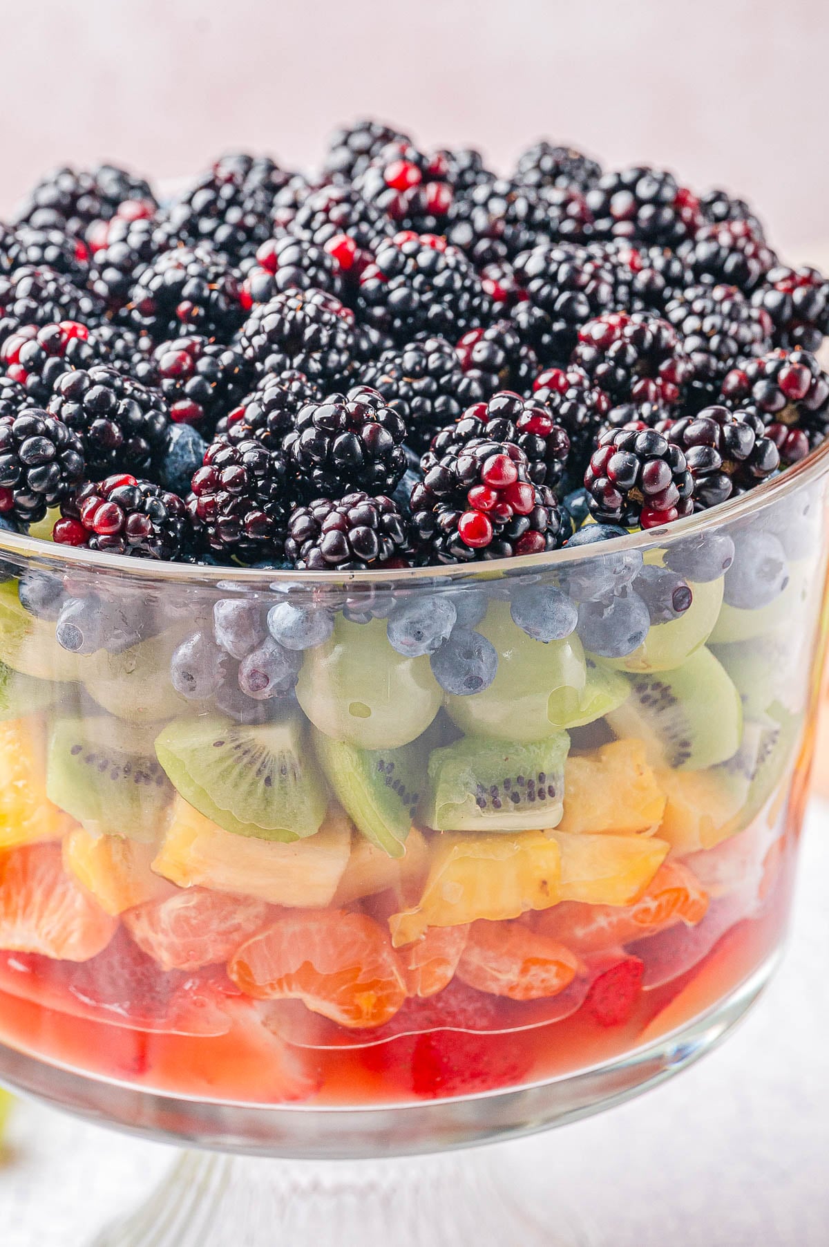 A glass trifle bowl filled with layered fresh fruit, including blackberries, blueberries, kiwi, pineapple, orange slices, and strawberries.