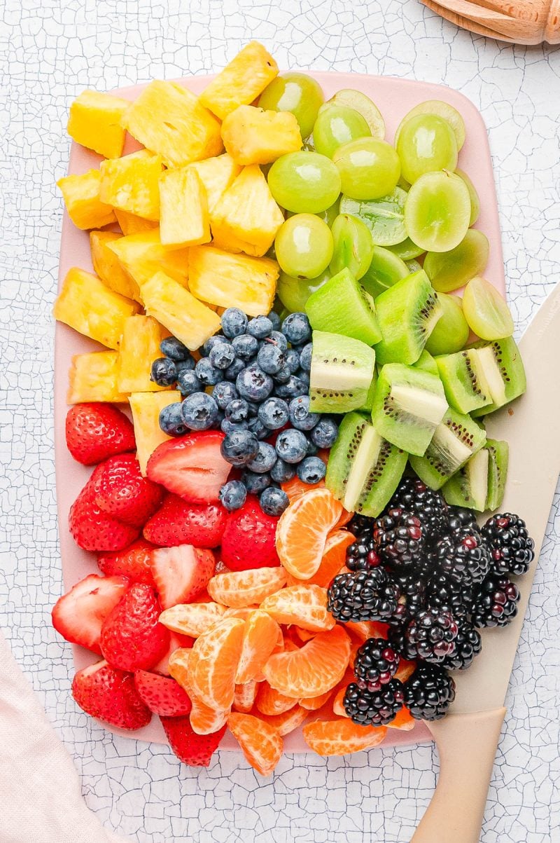 A cutting board with neatly arranged sections of pineapple, green grapes, kiwi, blueberries, strawberries, mandarin orange segments, and blackberries next to a knife.