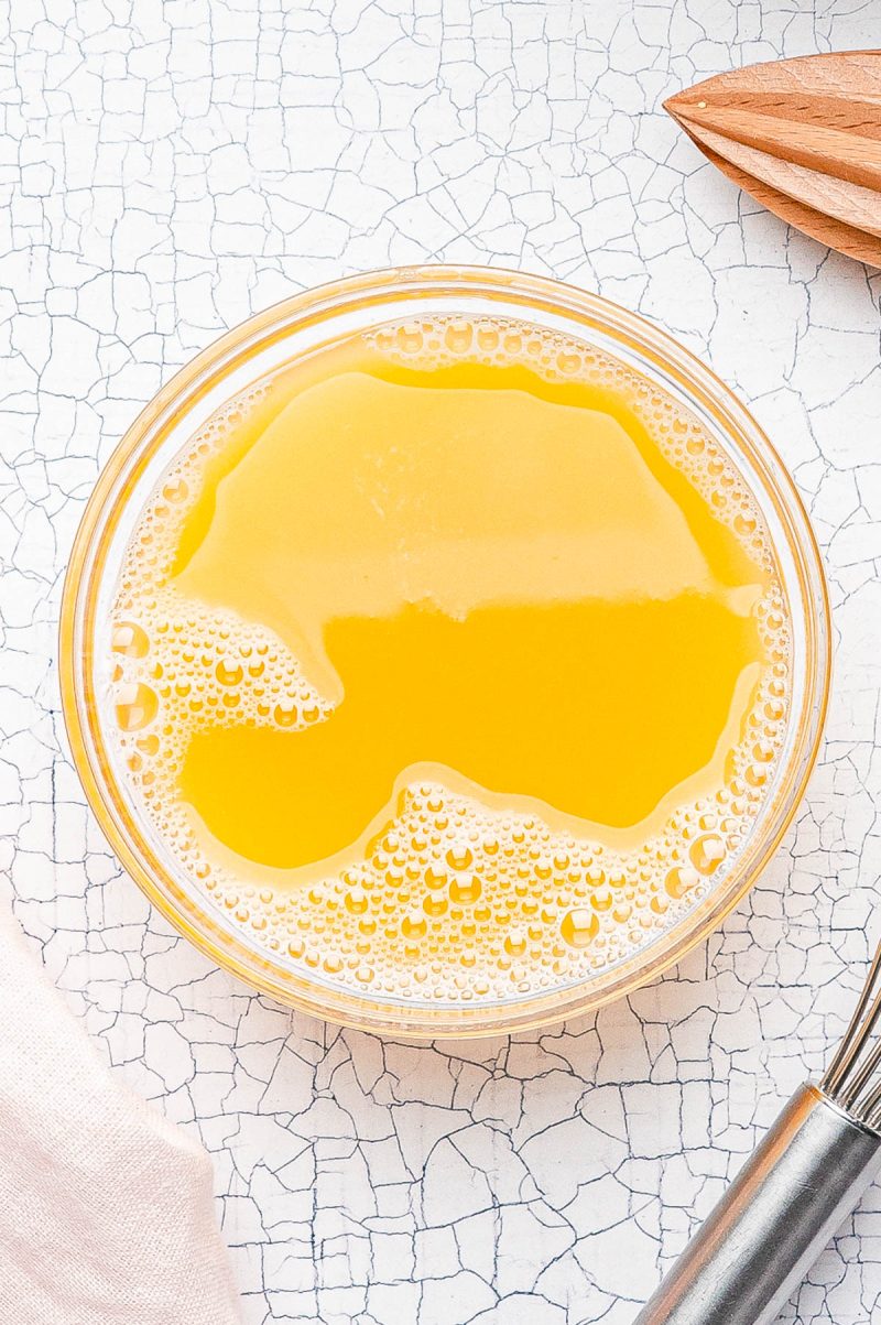 A clear glass bowl filled with yellow-orange liquid and bubbles sits on a white textured surface, next to a metal whisk and a wooden citrus reamer.