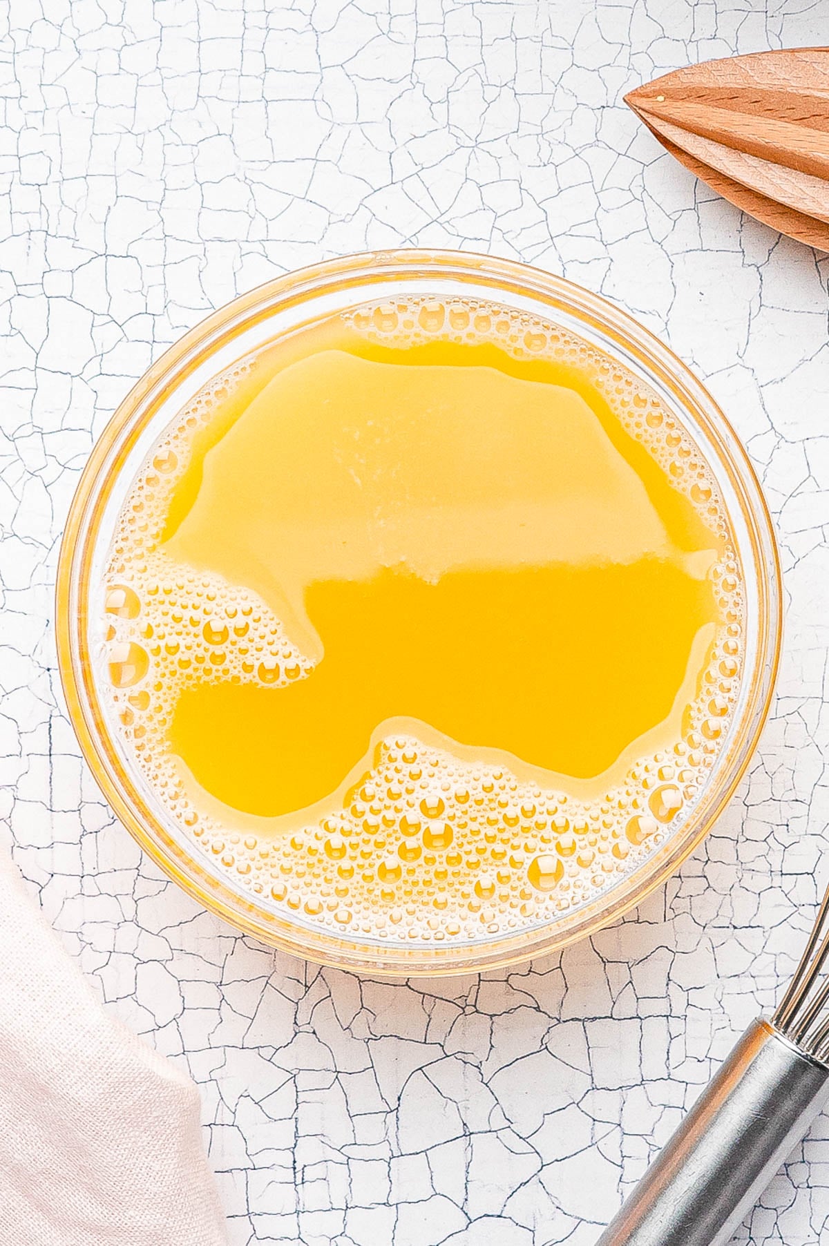 A clear glass bowl filled with yellow-orange liquid and bubbles sits on a white textured surface, next to a metal whisk and a wooden citrus reamer.