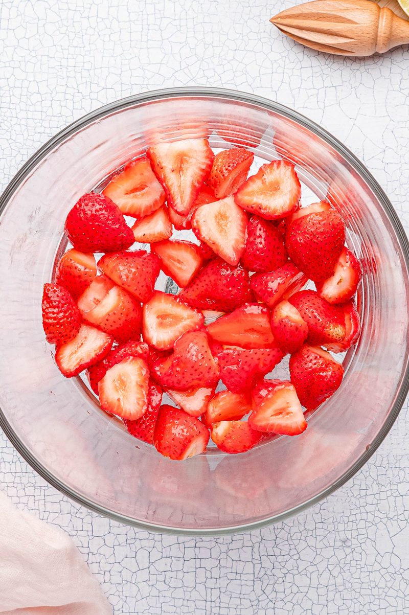 Halved strawberries in a clear glass bowl on a white textured surface, with a wooden citrus reamer and a light pink cloth nearby.