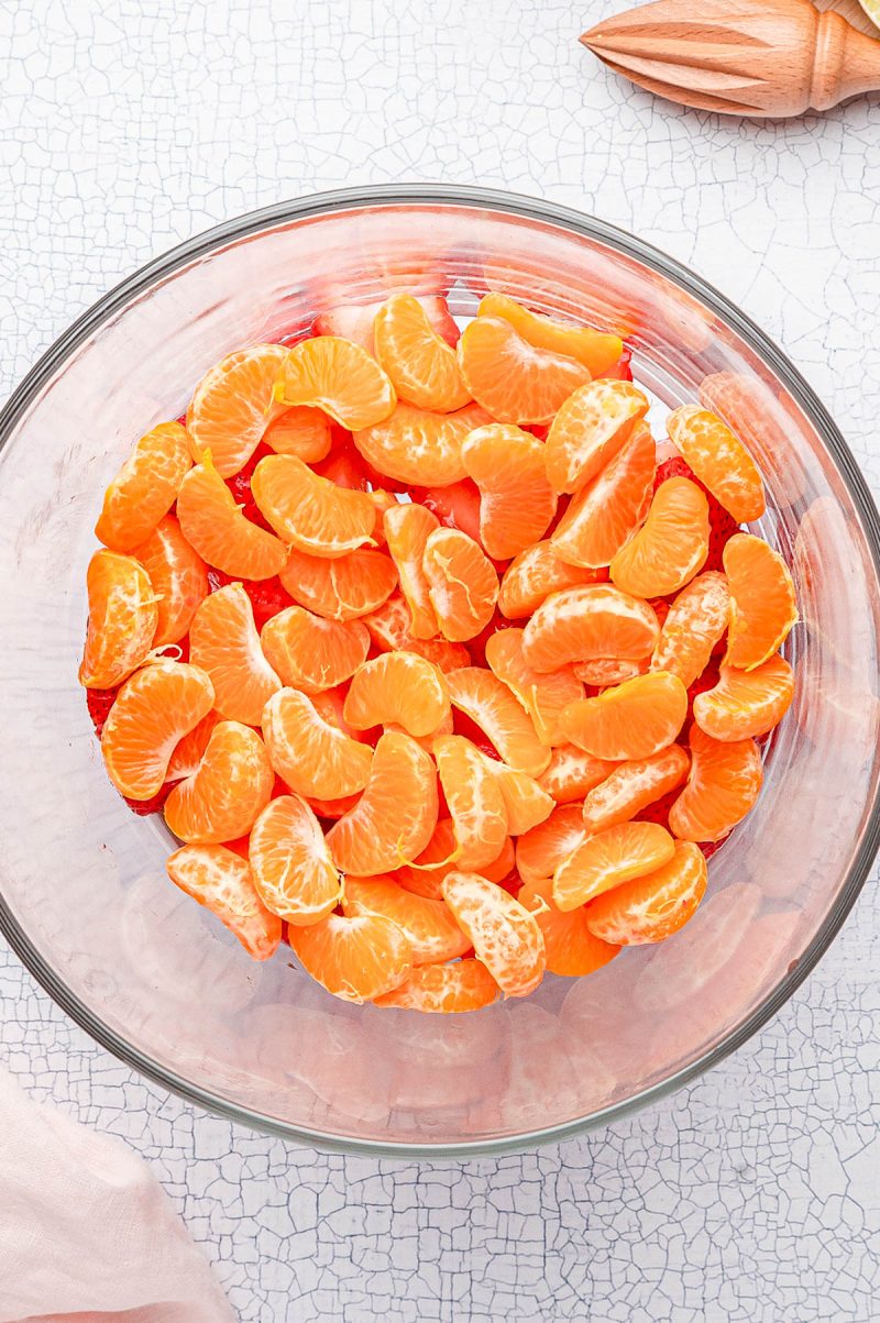 A glass bowl filled with peeled mandarin orange segments on a light textured surface, with a wooden citrus reamer nearby.