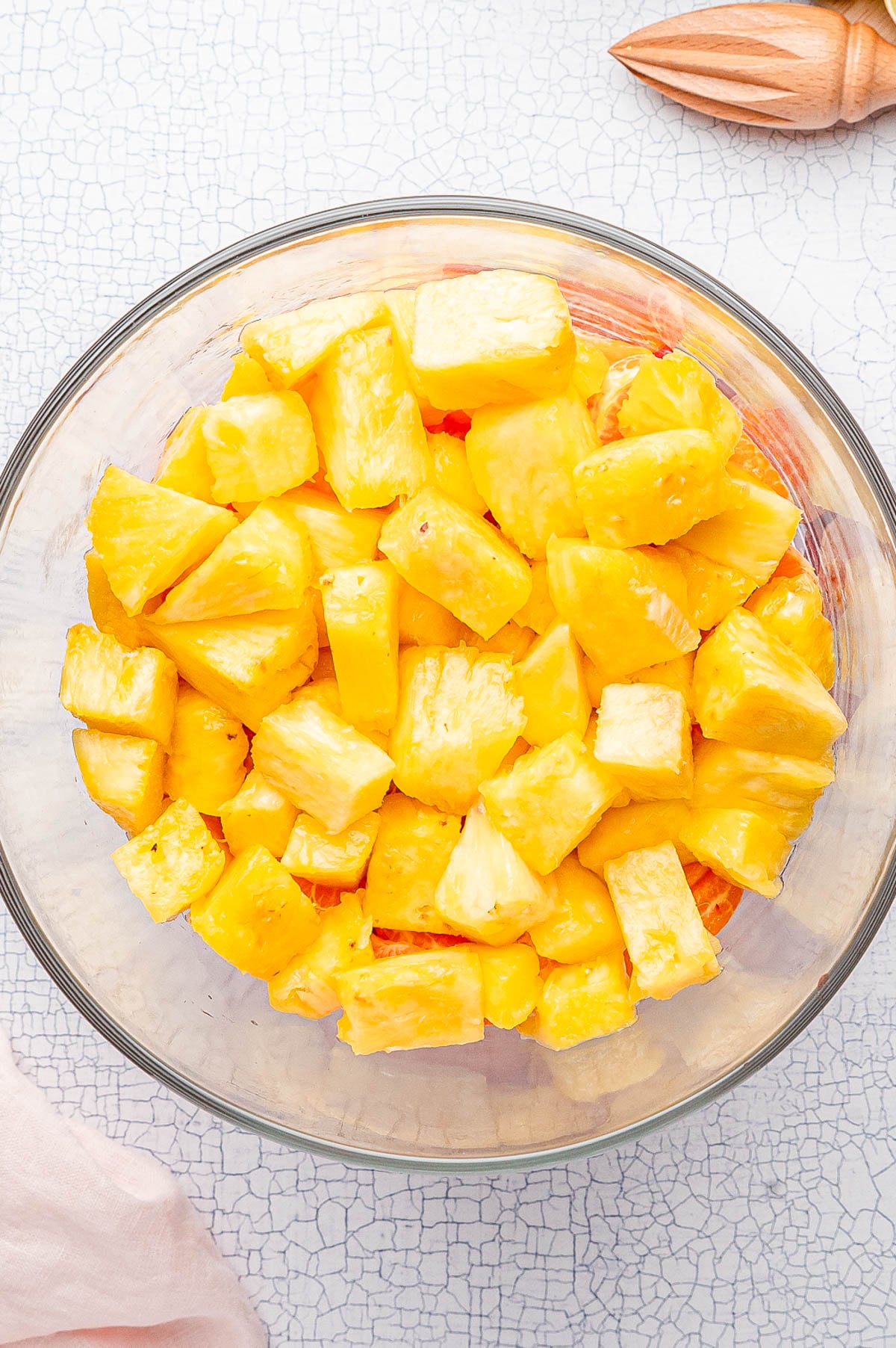 A clear glass bowl filled with freshly cut pineapple chunks sits on a white textured surface.
