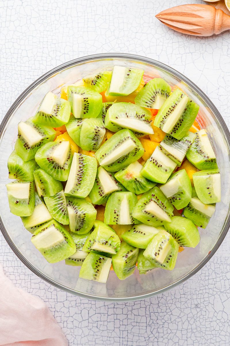 A glass bowl filled with chopped green kiwi pieces, placed on a light textured surface with a citrus juicer and pink cloth nearby.