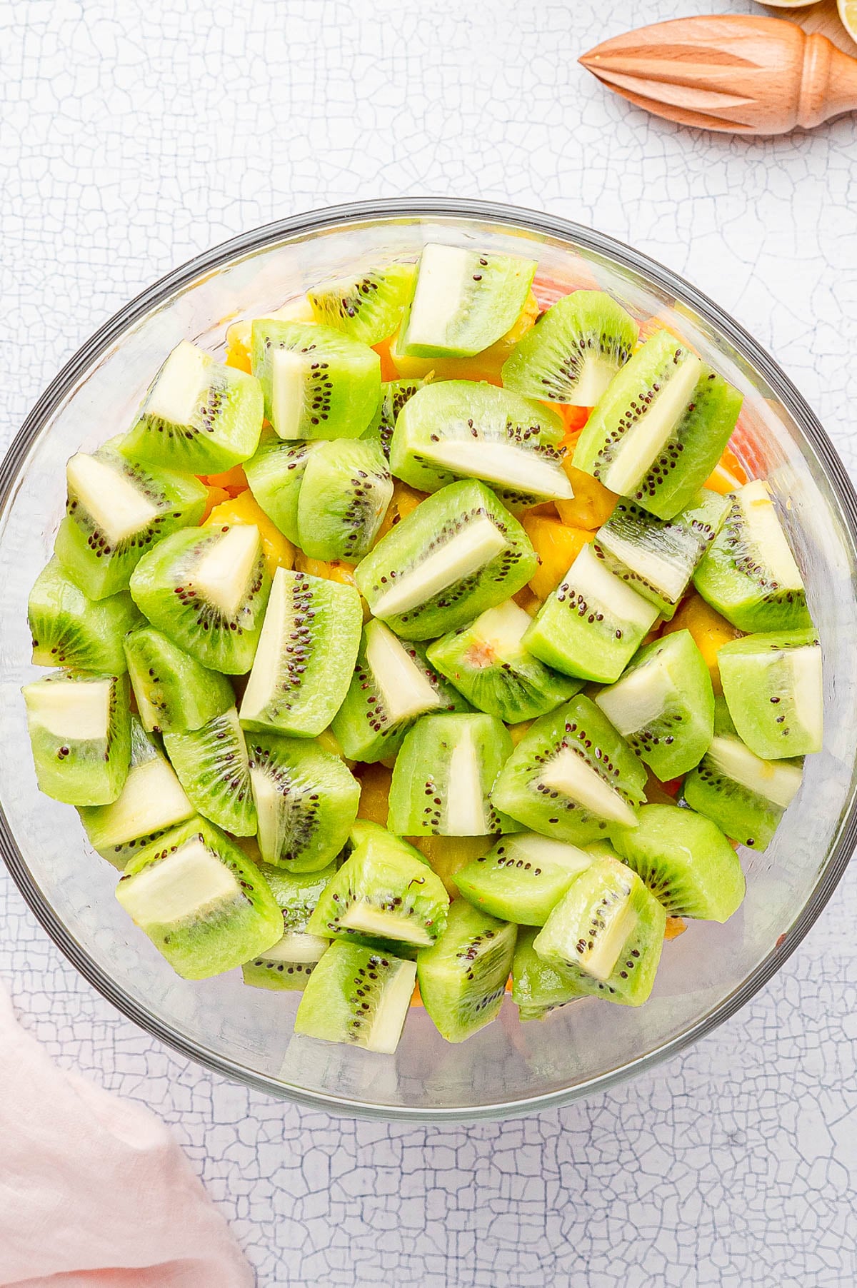 A glass bowl filled with chopped green kiwi pieces, placed on a light textured surface with a citrus juicer and pink cloth nearby.