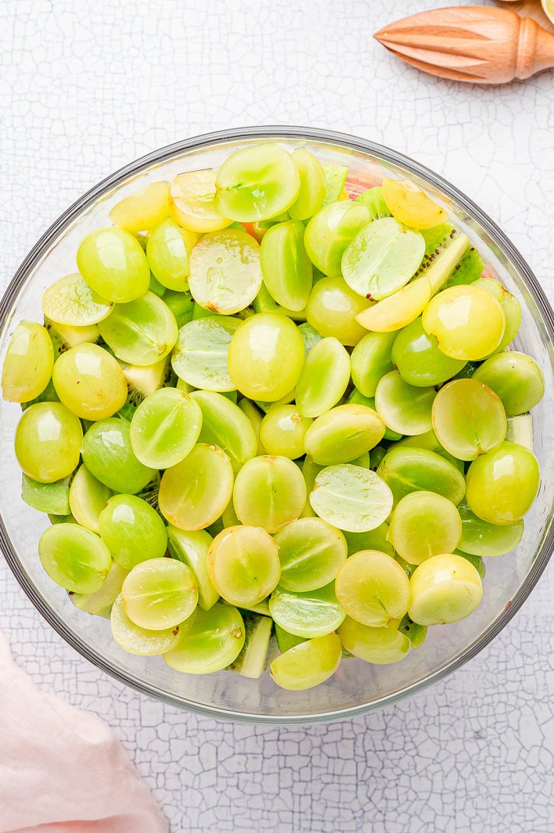 A glass bowl filled with halved green grapes on a light textured surface, with a wooden citrus juicer partially visible in the background.