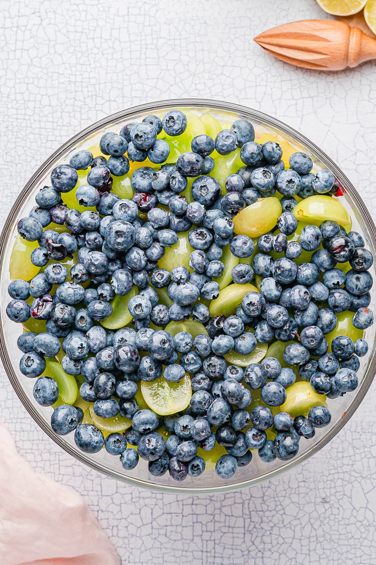 A clear bowl filled with green grapes and blueberries, viewed from above, on a light textured surface with a wooden citrus juicer nearby.