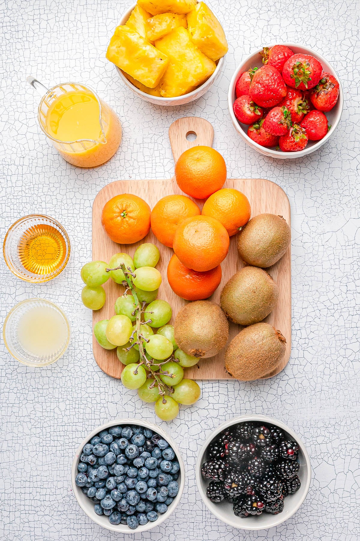 A variety of fresh fruits including tangerines, kiwis, green grapes, strawberries, pineapple, blueberries, and blackberries arranged on a table with small bowls of juice.
