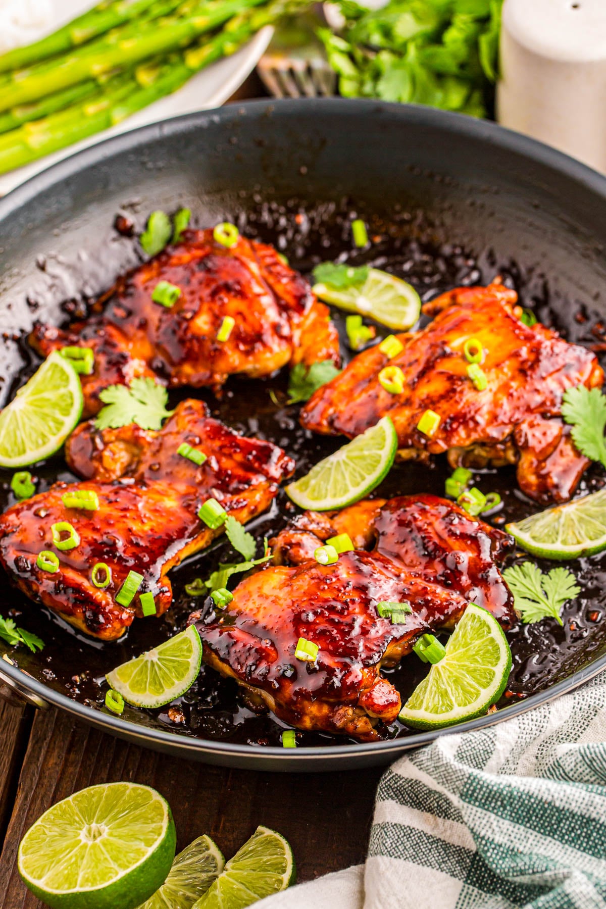 Chicken thighs glazed with sauce in a skillet, garnished with lime wedges, chopped green onions, and cilantro. Asparagus and fresh herbs are visible in the background.