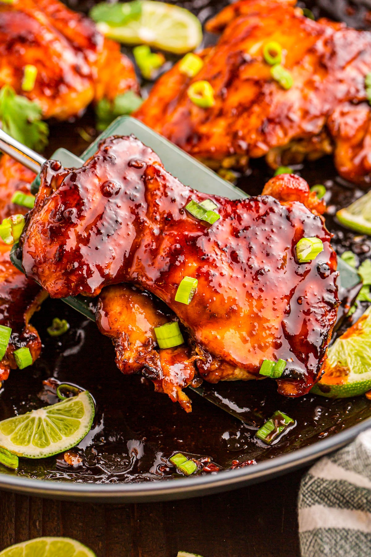 A glazed piece of cooked chicken topped with chopped green onions is being lifted from a pan with lime slices and more chicken pieces.