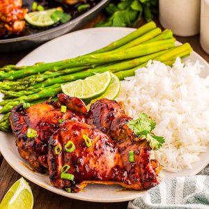 Plate of glazed chicken thighs with white rice, steamed asparagus, lime wedges, and green onion garnish on a table with a striped napkin and salt and pepper shakers.