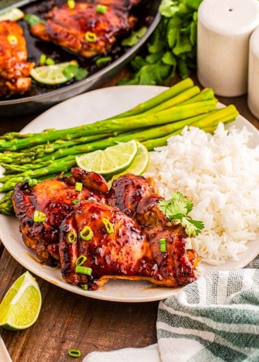 Plate of glazed chicken thighs with white rice, steamed asparagus, lime wedges, and green onion garnish on a table with a striped napkin and salt and pepper shakers.