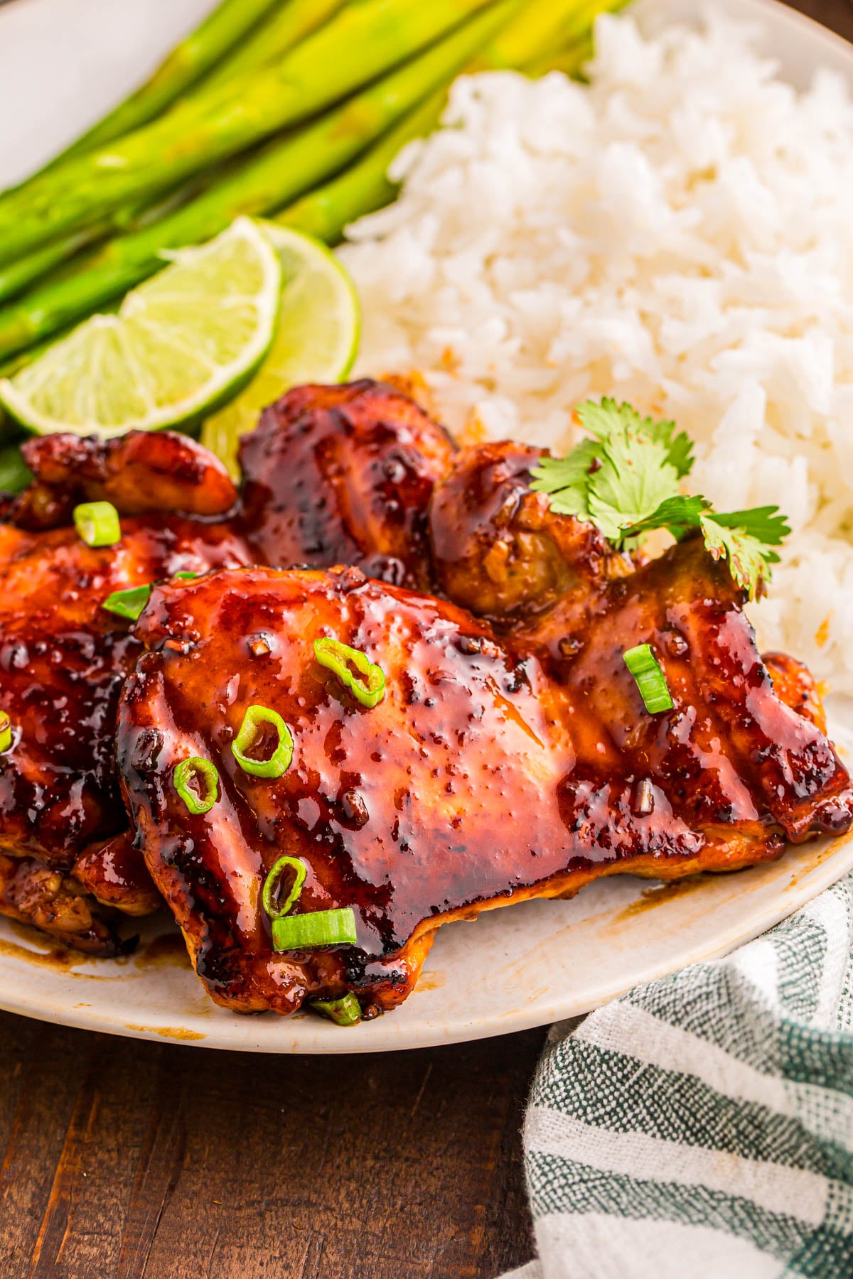 Plate of glazed chicken thighs topped with sliced green onions, served with white rice, asparagus spears, lime wedges, and a garnish of cilantro.