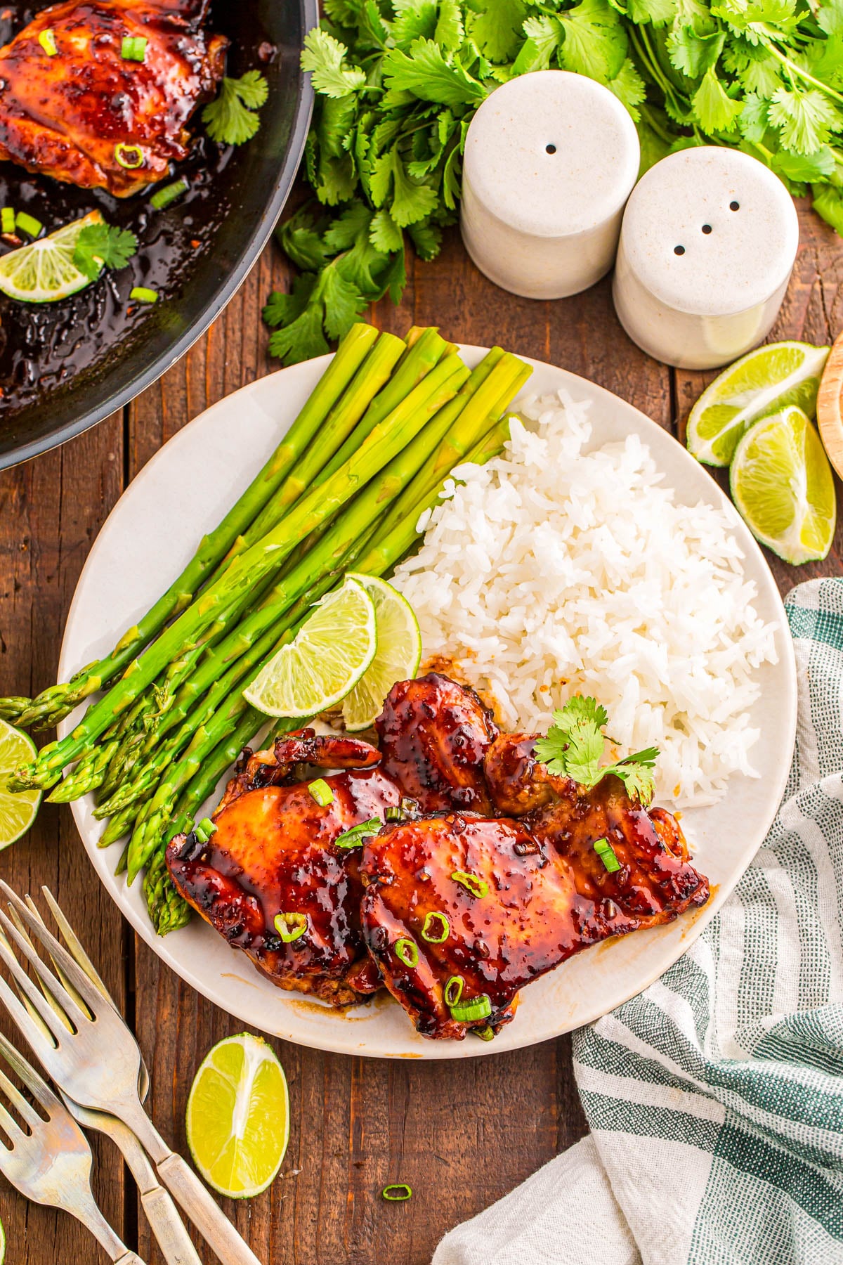 A plate with glazed chicken thighs, white rice, and steamed asparagus, garnished with lime wedges and cilantro, next to a pan of chicken and fresh herbs on a wooden table.