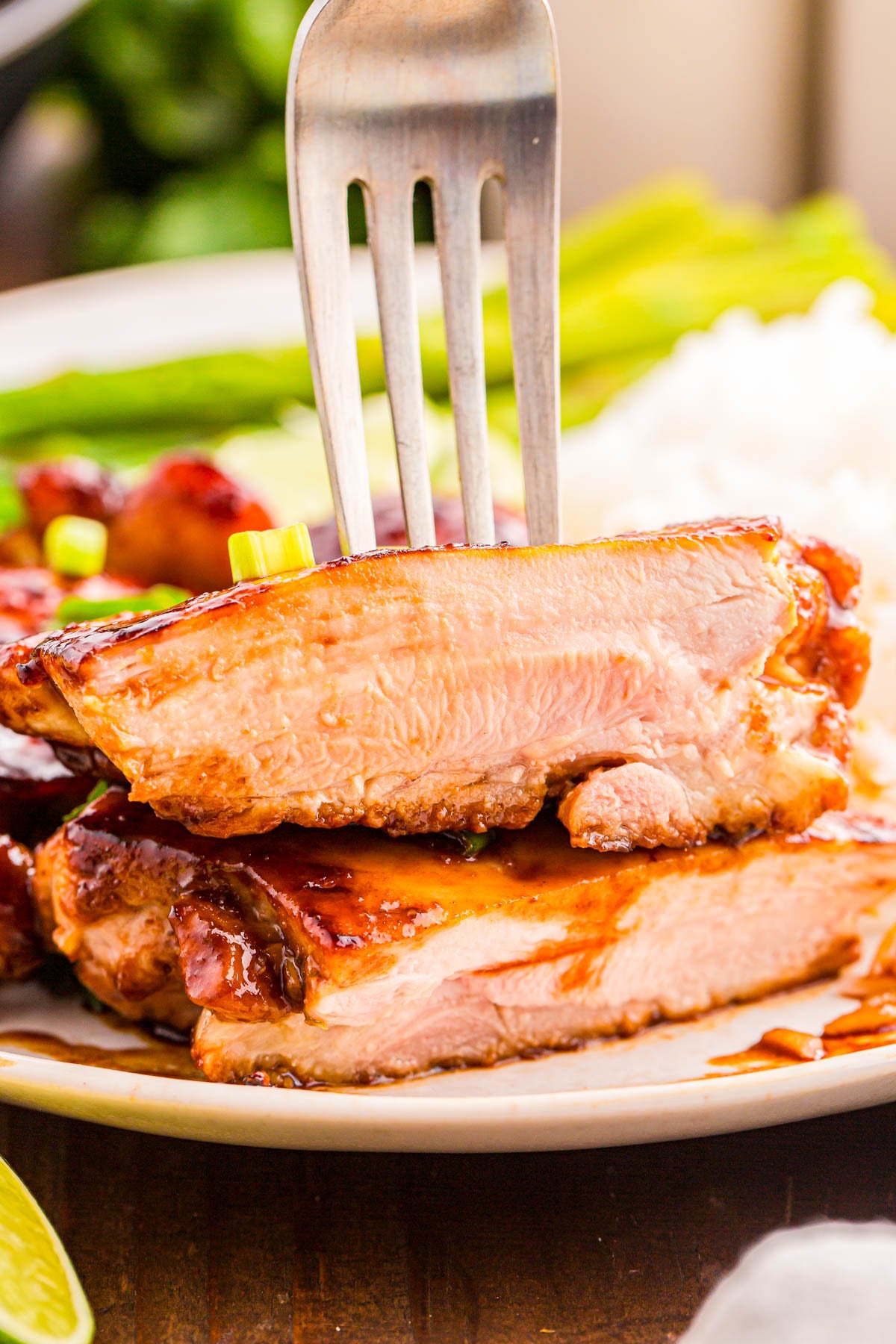 Two pieces of cooked chicken stacked on a plate, pierced by a fork, with rice and green vegetables in the background.