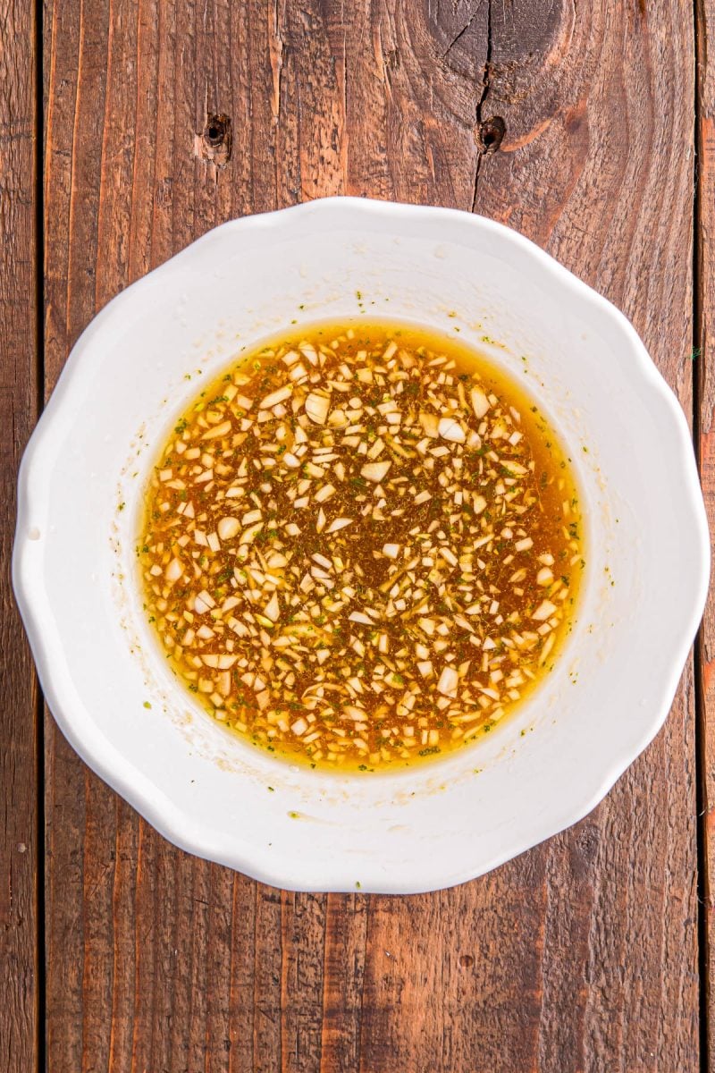 White bowl containing a mixture of finely chopped garlic and herbs in a light brown liquid, placed on a wooden surface.