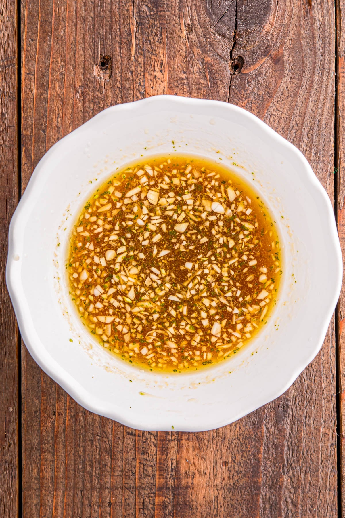 White bowl containing a mixture of finely chopped garlic and herbs in a light brown liquid, placed on a wooden surface.