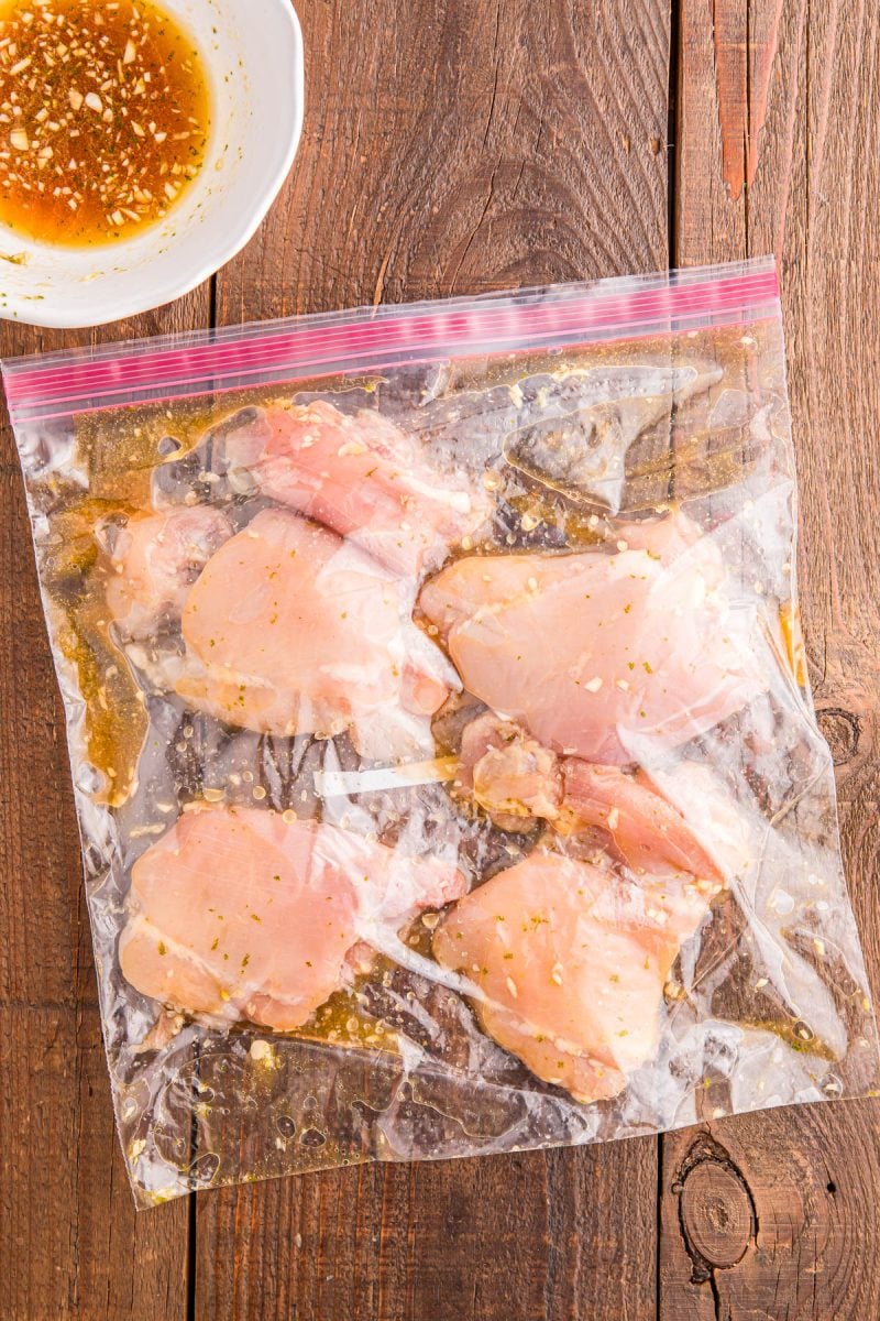 Raw chicken pieces marinating in a resealable plastic bag with a visible marinade mixture; a bowl with leftover marinade sits nearby on a wooden surface.