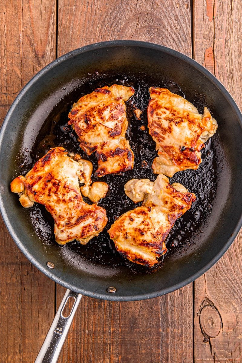 Four browned pieces of chicken cooking in a black frying pan on a wooden surface.