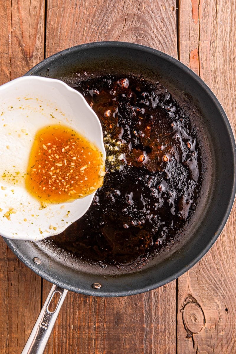 A white bowl pours a light brown liquid with seasonings into a blackened skillet on a wooden surface.