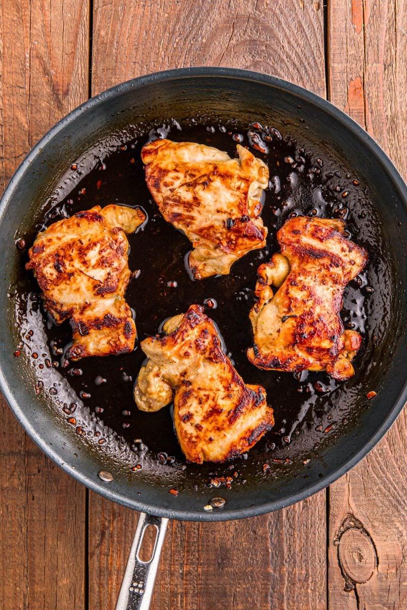 Four pieces of browned chicken cooking in a black frying pan with sauce, set on a wooden surface.