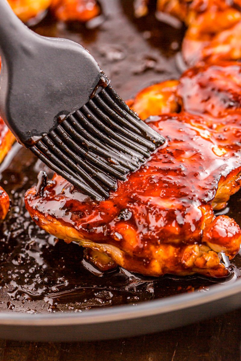 A close-up of a silicone brush coating cooked chicken pieces with barbecue sauce in a pan.