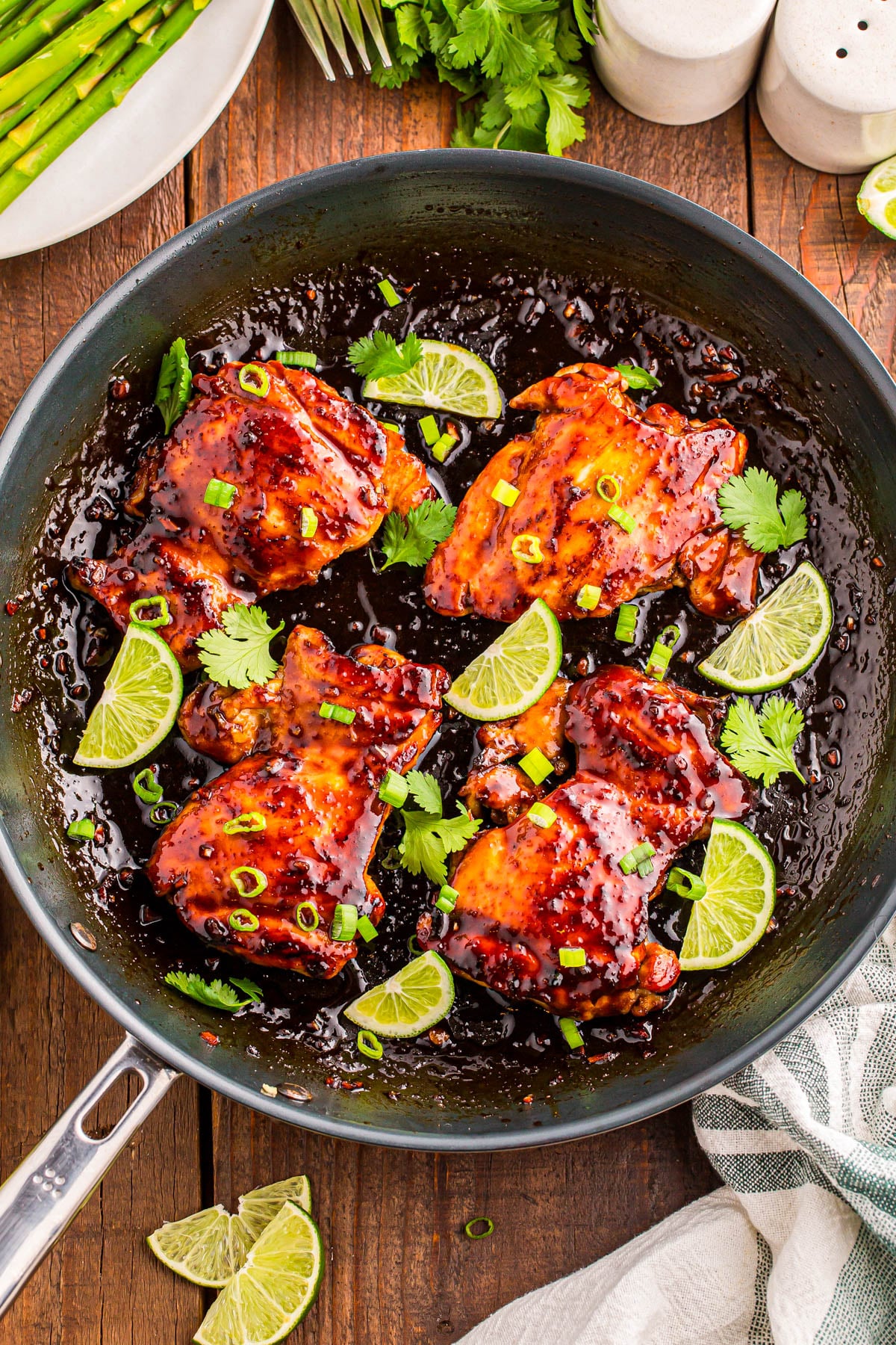 A skillet with glazed chicken thighs garnished with lime wedges, cilantro, and green onions, sitting on a wooden table with asparagus and seasonings nearby.