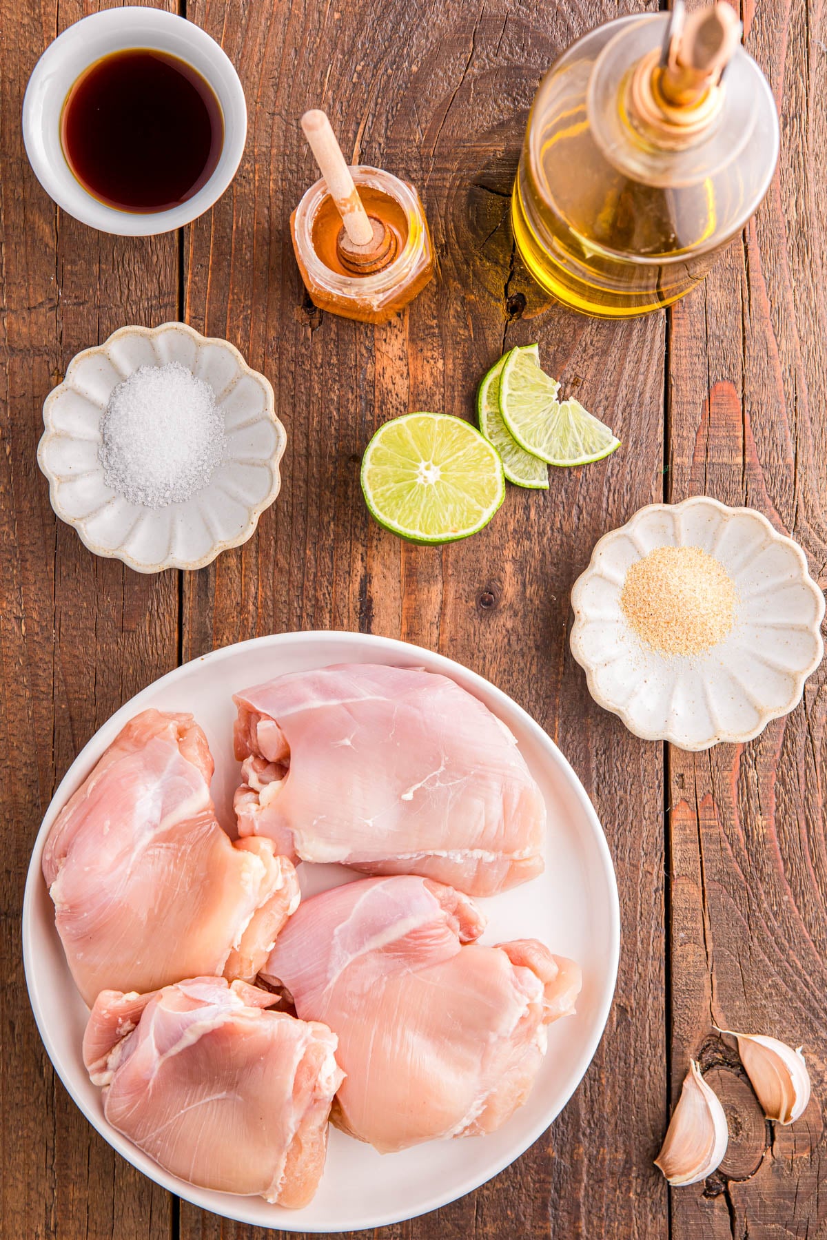 Raw chicken thighs on a plate surrounded by small bowls of salt, garlic powder, soy sauce, honey, sliced lime, olive oil, and garlic cloves on a wooden surface.