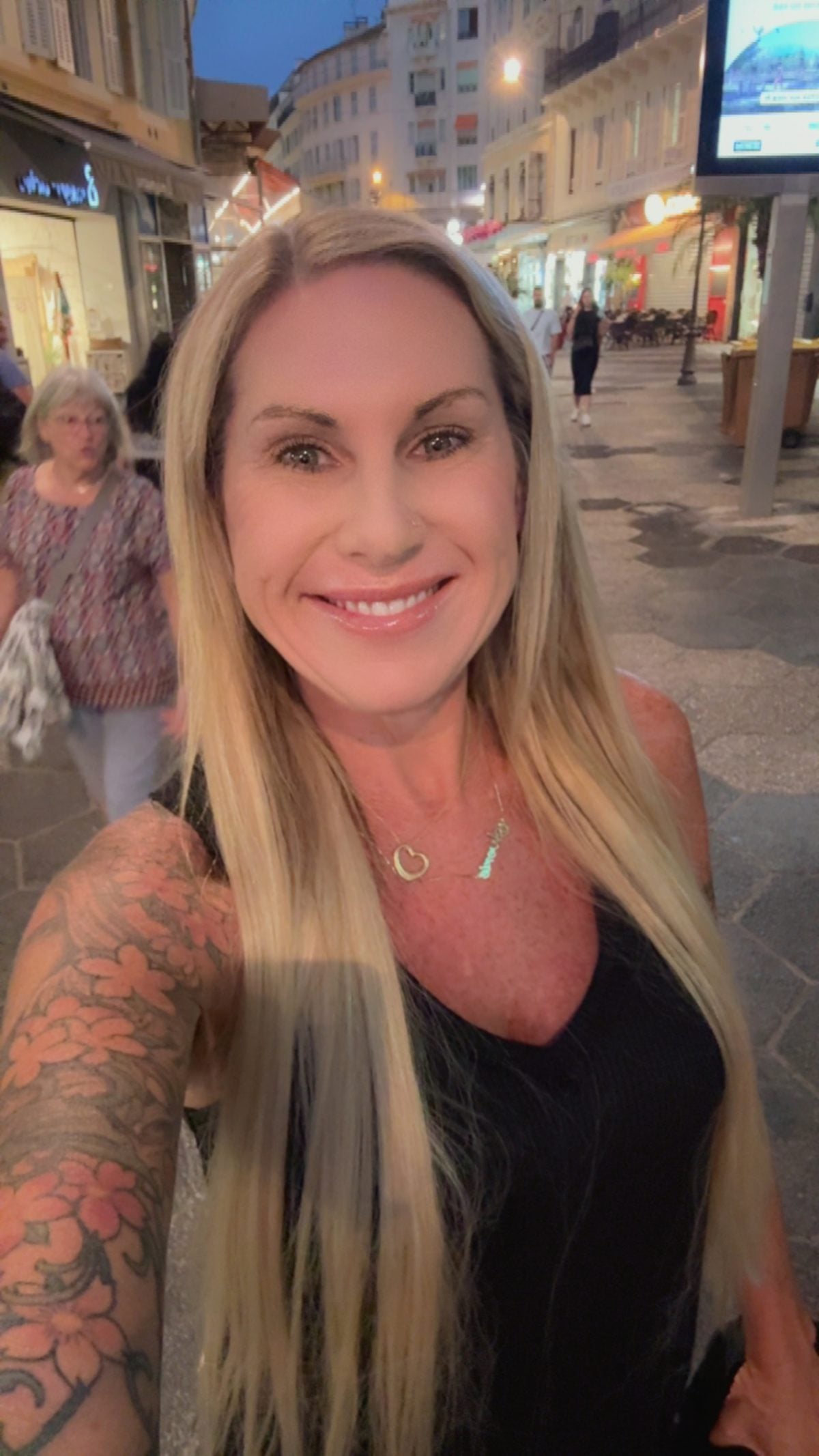 Woman with long blonde hair smiles for a selfie on a city street at dusk; people and shops are visible in the background.