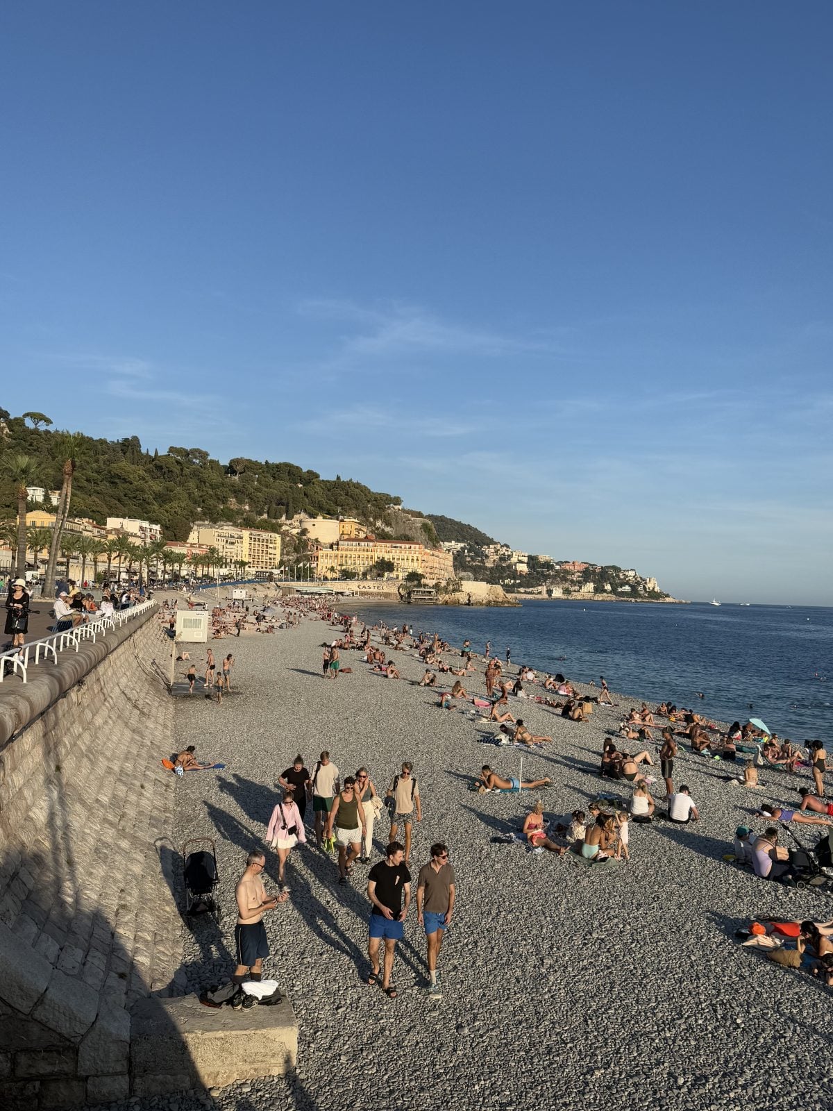People are relaxing and walking on a pebbly beach by the sea, with buildings and greenery visible in the background under a clear blue sky.