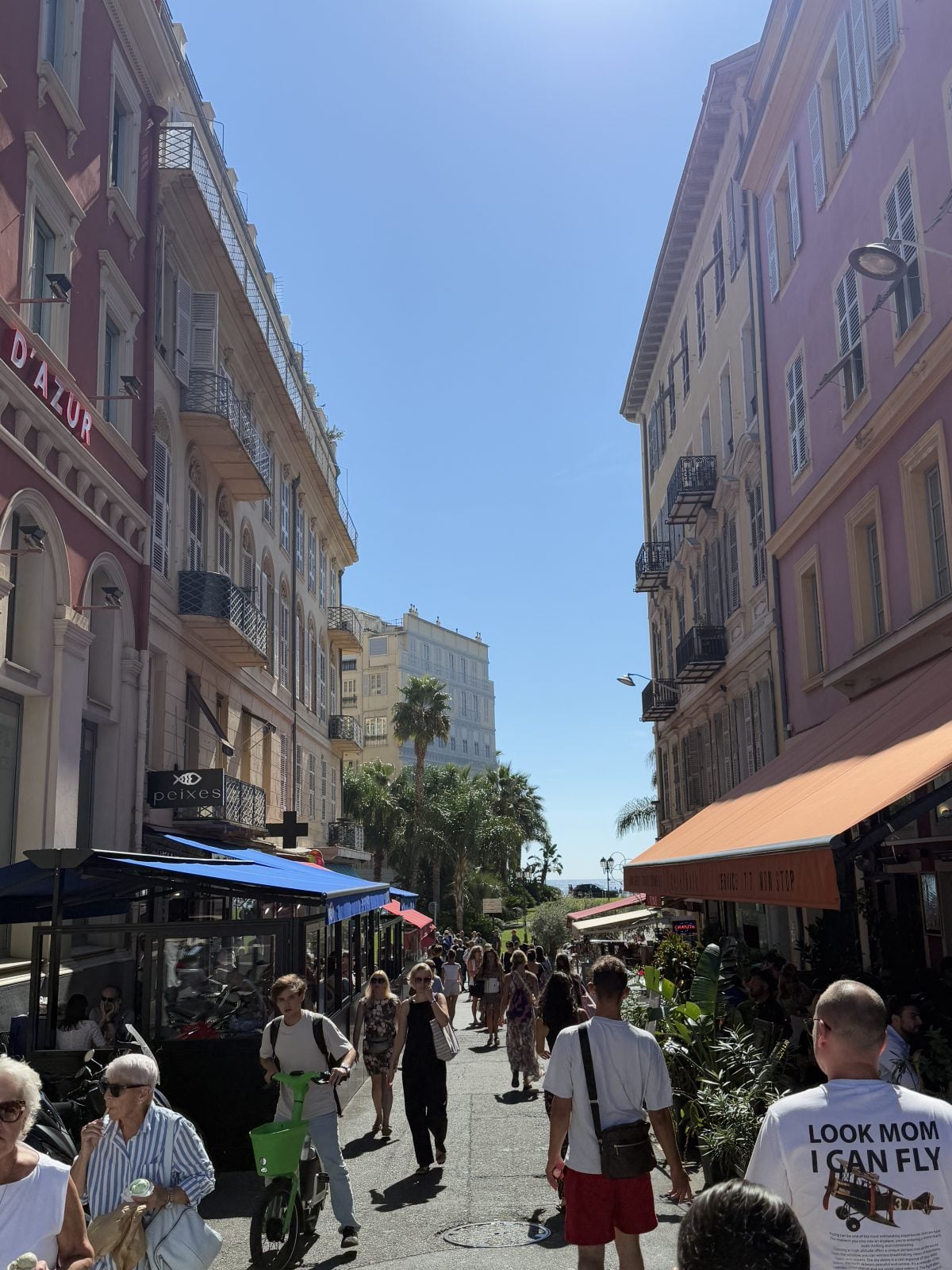A busy pedestrian street lined with cafes and shops on a sunny day, people walking and socializing, with palm trees and a glimpse of the sea in the background.