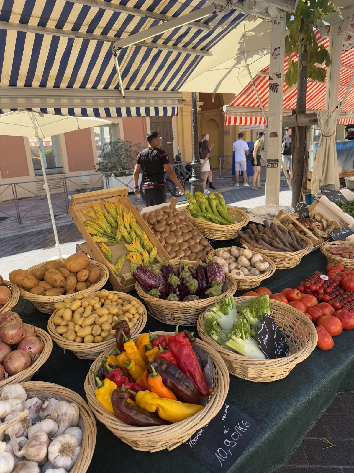 A market stall displays baskets of assorted fresh vegetables, including peppers, potatoes, eggplants, garlic, and tomatoes, under a striped canopy.