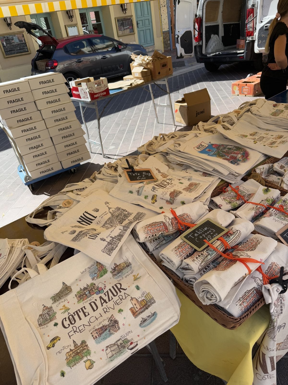 Tote bags with Cote d'Azur designs are displayed on a market stall, with boxes labeled "fragile" stacked nearby and a van parked in the background.