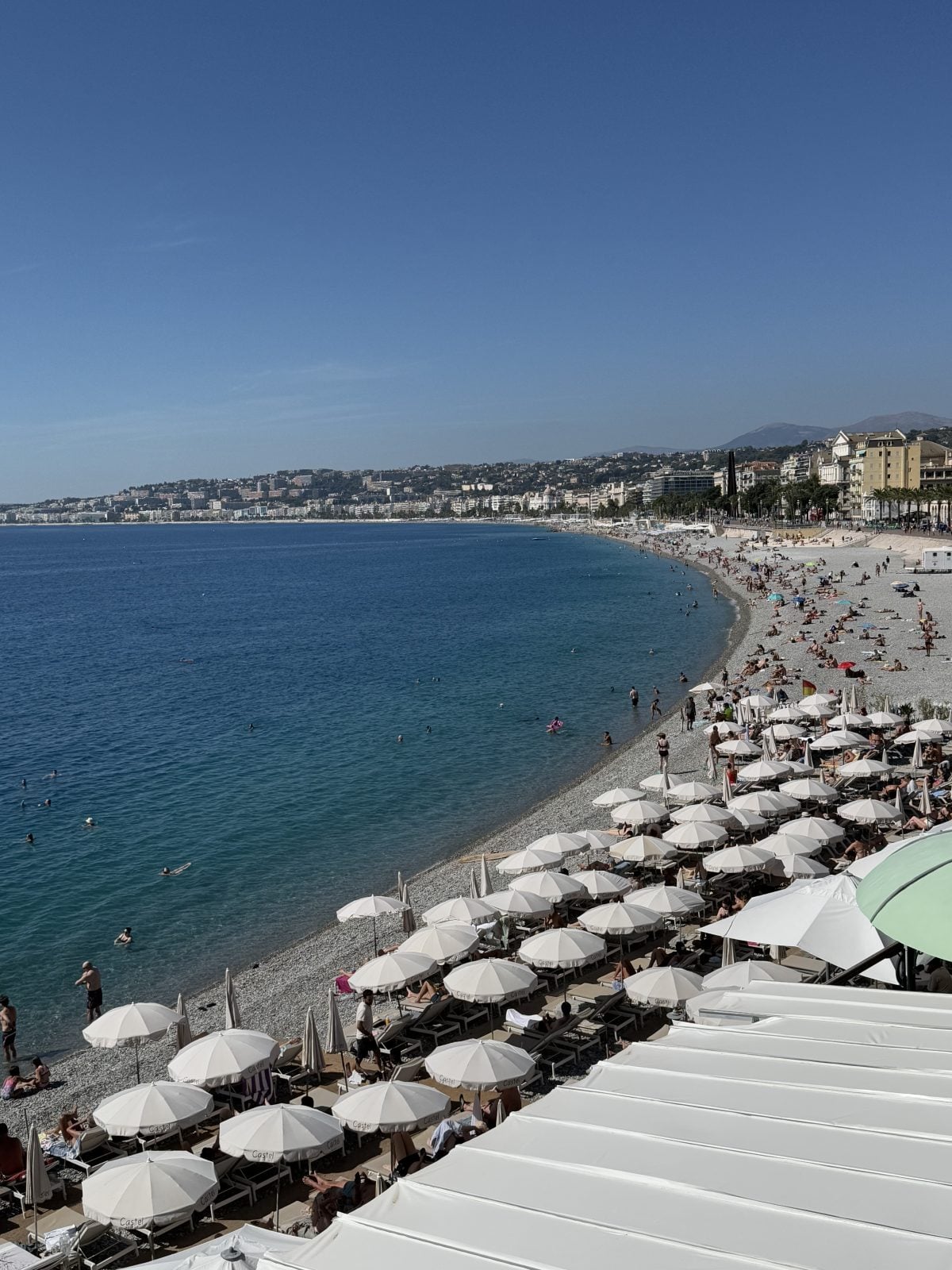 A crowded pebbly beach with rows of white umbrellas and sun loungers, people swimming in the blue sea, and a city skyline in the background under a clear sky.