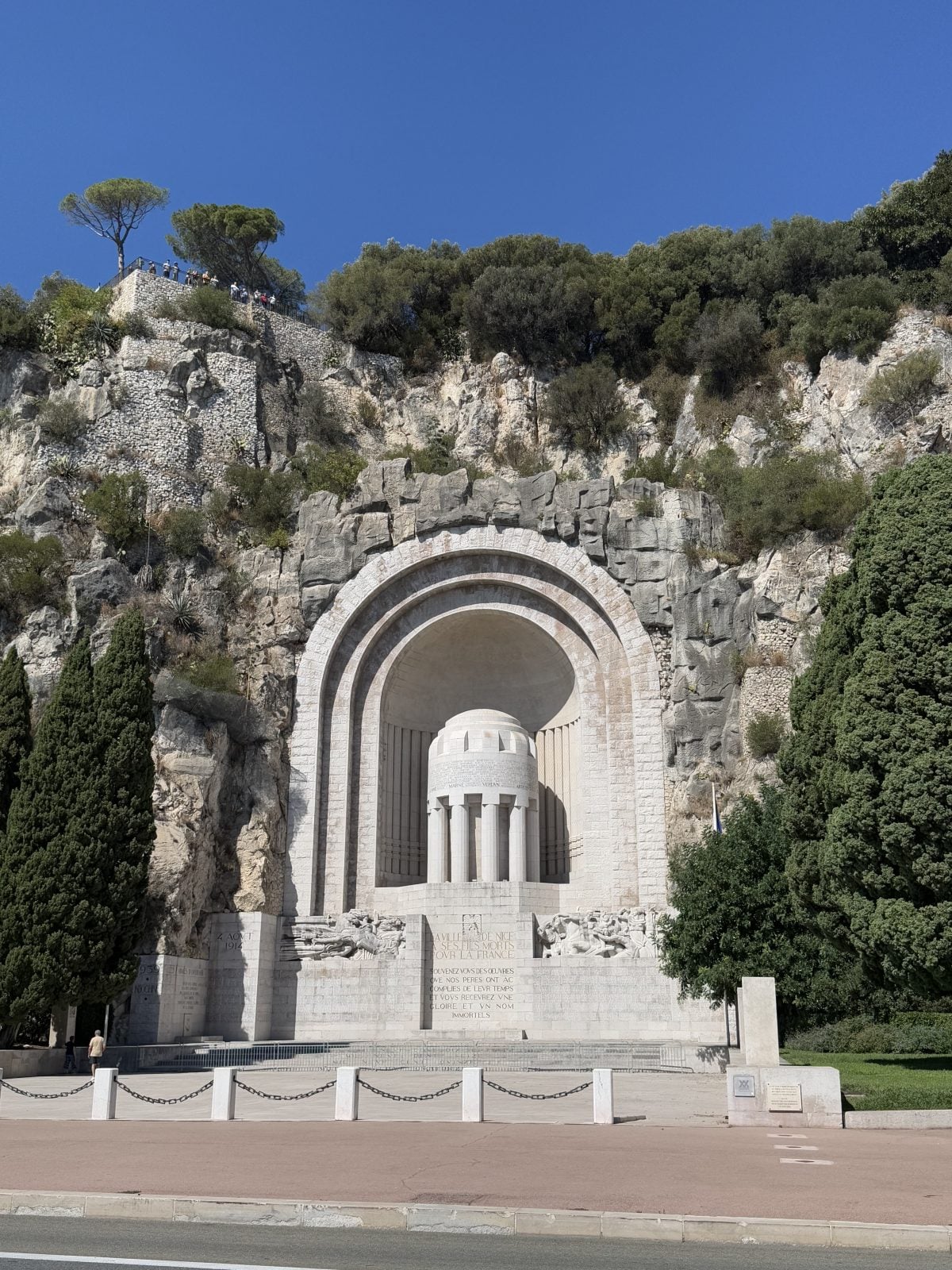 A large stone war memorial with arched entrance and domed structure is built into a rocky hillside, surrounded by trees under a clear blue sky.