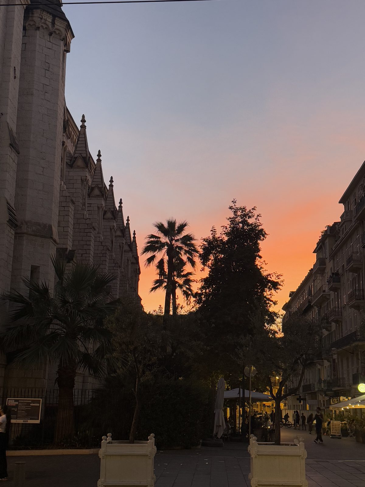 City street scene at sunset with a church on the left, palm trees and other foliage, and buildings with lit-up shops and cafes on the right.