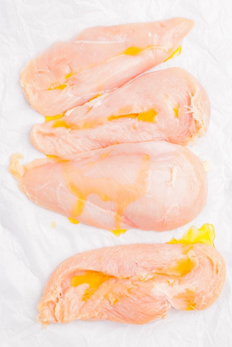 Four raw chicken breasts placed on white parchment paper, drizzled lightly with oil.