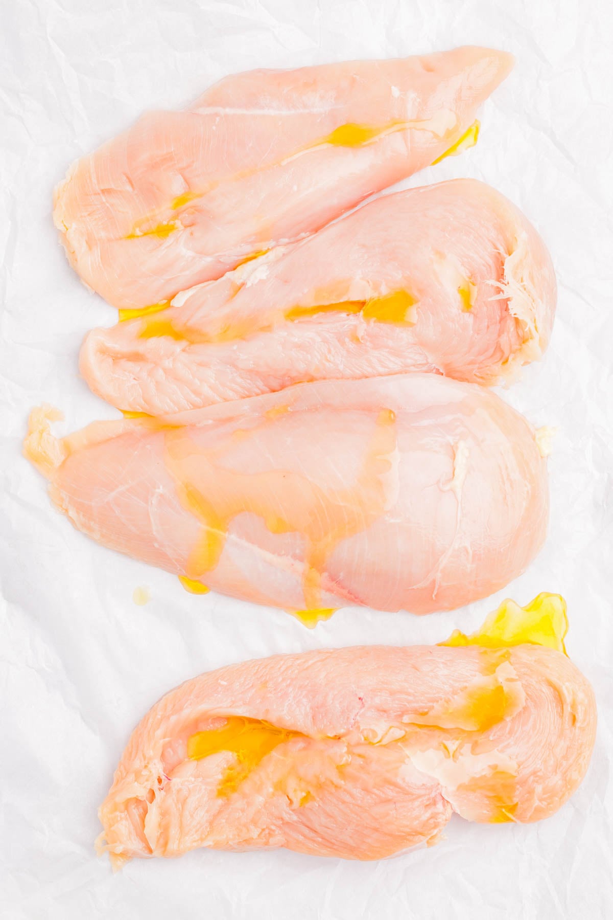 Four raw chicken breasts placed on white parchment paper, drizzled lightly with oil.